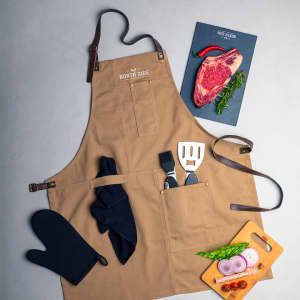 A tan apron with pockets, accompanied by kitchen utensils, an oven mitt, and fresh ingredients.