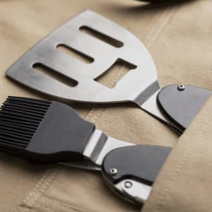 A beige apron with a metal spatula and a brush tucked into its pocket, featuring a logo.