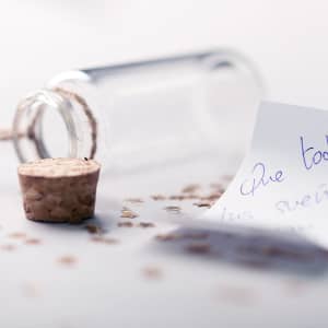A clear glass wish bottle with a cork stopper, surrounded by scattered glitter and a handwritten note.