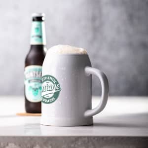 A white beer mug with a frothy top, featuring a logo and beside a beer bottle on a surface.