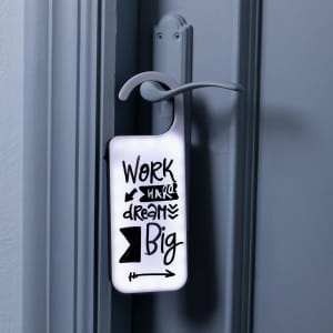 A decorative door hanger in white with black text and designs, featuring motivational phrases and a hook for hanging.