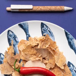 A cork-textured pen with a silver clip lies next to a plate of fish-patterned wood shavings and a red chilli.