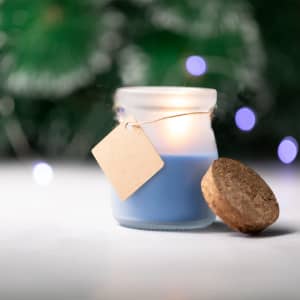 A blue aromatic candle in a glass jar with a cork lid and a blank tag, surrounded by soft, out-of-focus lights.