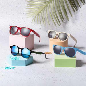 Five pairs of sunglasses in red, white, black, green, and blue displayed on pastel geometric blocks with greenery.
