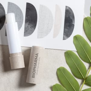 A lip balm in a sleek white tube with a cardboard base, surrounded by leafy green plants and textured artistic prints.