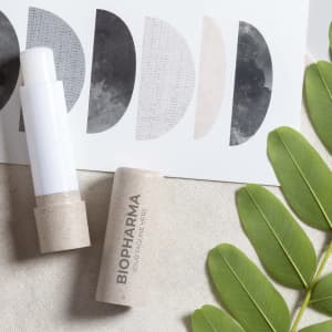 A white and natural-coloured lip balm tube beside a patterned card and green leaves on a textured surface.