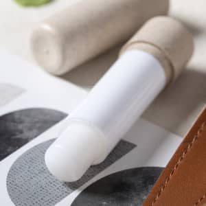 A white lip balm in a cylindrical container with a natural-toned cap, beside textured paper and a leather item.
