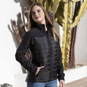 A woman stands wearing a black soft shell jacket with a zip front and padded sections, alongside cacti in the background.