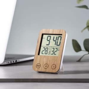 A wooden-framed weather station with a digital display, showing time and temperature, placed on a desk near a laptop.