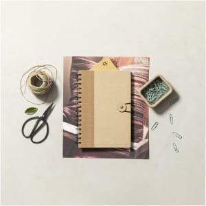 A brown spiral-bound notebook with a button closure, surrounded by scissors, twine, and stationery clips on a textured background.
