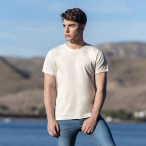 An organic natural adult T-shirt in cream, worn by a model outdoors with a scenic background. The shirt features a logo.