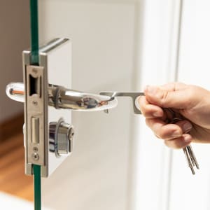 A metallic keyring tool being used to open a door lock, held by a hand. The tool is silver and sleek in design.