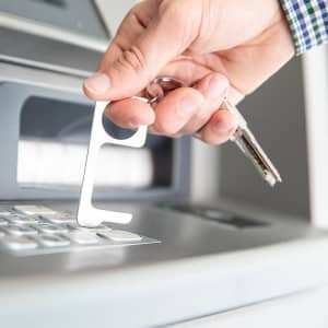 A silver keyring tool is being used to press buttons on an ATM, held by a hand.