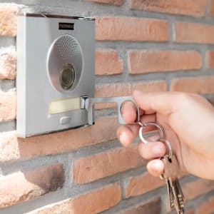 A keyring tool in silver, being used to press a button on a wall-mounted intercom. Keys attached to the ring are visible.