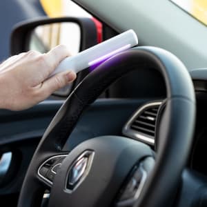 A white Klas UV Sterilizer Lamp being used on a car steering wheel. The lamp emits a purple light.