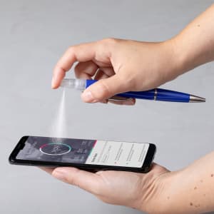 A blue spray pen in use, releasing a fine mist above a smartphone on a grey surface. The pen has a logo.