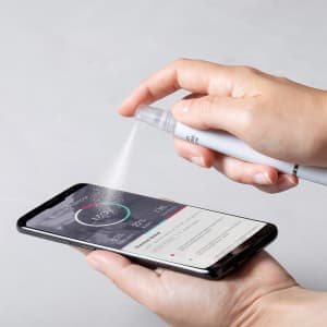A white multifunction pen sprays a fine mist towards a smartphone screen, showcasing its antibacterial feature.