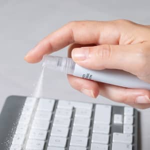 A hand holding a spray pen releasing particles onto a computer keyboard, featuring a sleek design in white.