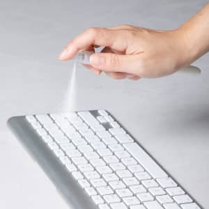 A hand holds a spray pen, dispersing a fine mist above a silver and white keyboard. The pen has a clear body.