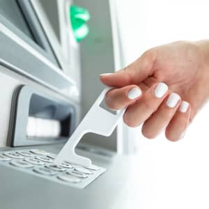 A white antibacterial keyring tool interacts with an ATM keypad. The tool is held by a hand with neatly painted nails.