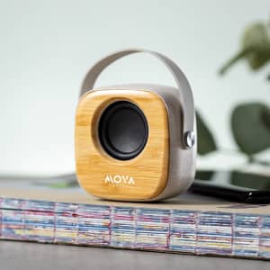 A square speaker with a bamboo finish and a grey body, featuring a carry handle, placed on a stack of magazines.