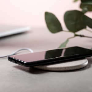 A wireless charger in a round shape with a phone resting on it, set against a neutral background and greenery.