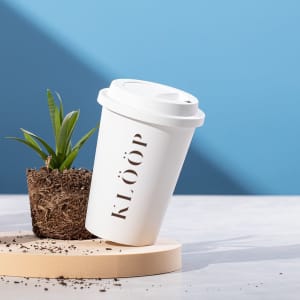A white plastic cup with a lid, leaning beside a small potted plant on a wooden surface. The cup features a logo.