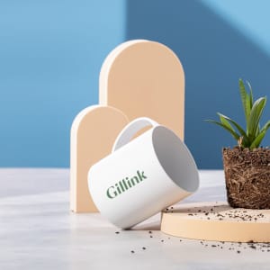 A white coffee mug with a handle, featuring a logo, positioned beside geometric shapes and a potted plant.
