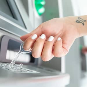 A hand holding a transparent antimicrobial keyring placed over an ATM keypad, with white nails and a tattoo visible.