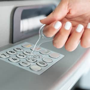 A transparent keyring tool is being used to press buttons on an ATM keypad, featuring a hand with white nail polish.