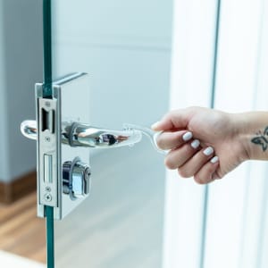A hand using a transparent, plastic anti-contact tool to open a glass door handle. The tool is designed for hygienic access.