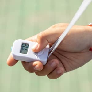 A white digital tally counter held in a hand, featuring a clear display and a strap.