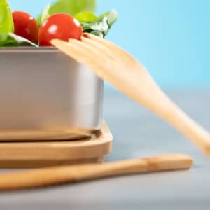 A stainless steel lunch box with a wooden serving fork, containing fresh spinach and cherry tomatoes on a bamboo base.
