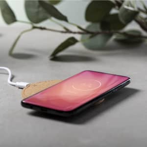 A wireless charger in a round, natural finish with a smartphone placed on top, surrounded by greenery.