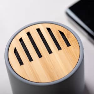 A grey speaker with a bamboo top featuring slotted openings, placed beside a smartphone.