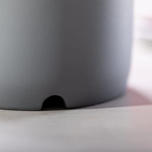 A grey speaker with a smooth texture and a small opening at the base, accompanied by a blurred background.