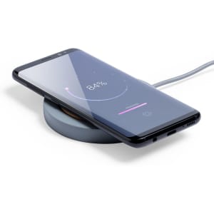 A wireless charger in grey with a smartphone on it, displaying a charging indicator on the screen.