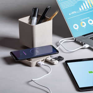 A beige multifunction pencil holder containing pens and a paintbrush, with a smartphone and charging cables nearby.