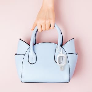 A light blue handbag with a hand sanitizer attached, set against a pink background.