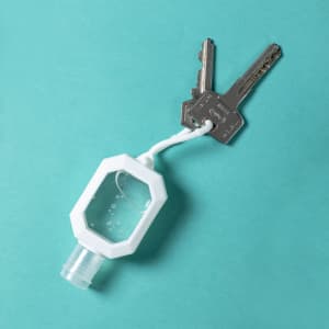 A clear hand sanitiser bottle with a white cap, attached to two metal keys, on a teal background.