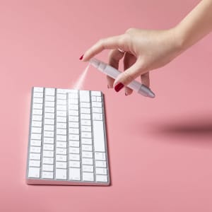 A hand holding a spray pen above a white keyboard, with a pink background. The pen has a sleek design and a logo.