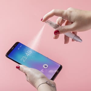 A person holding a white multifunction antibacterial pen, spraying mist onto a smartphone screen against a pink background.