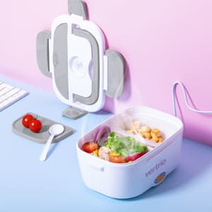 A white electric lunch box with compartments, containing vegetables and cubes of food, next to a plate of cherry tomatoes.