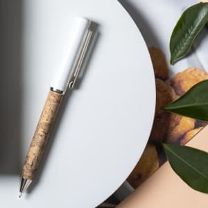 A metal pen with a cork body and white barrel, placed on a light grey surface with surrounding greenery.