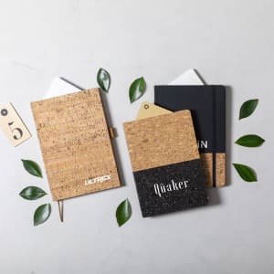 Three notebooks with cork and black covers placed among green leaves on a light background. One notebook has a logo.