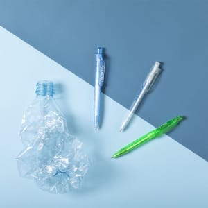 A selection of plastic pens in blue, clear, and green, alongside a crumpled plastic bottle on a blue and pale blue background.