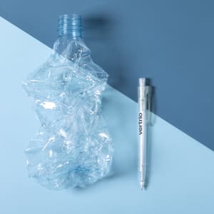 A crumpled plastic drink bottle and a clear plastic pen are positioned on a two-tone blue background.
