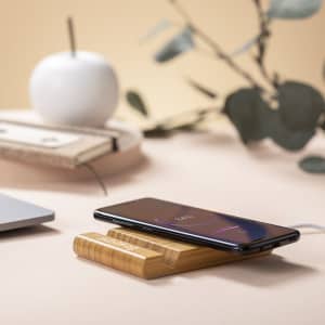 A wooden wireless charger in natural tones, with a smartphone resting on it, surrounded by green foliage and a white apple.