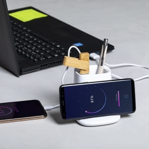 A white multifunction pencil holder with wooden clips, a pen, and a smartphone charging on a wireless charger beside a laptop.