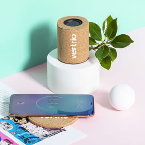 A cork speaker in a light blue setting with a smartphone and a small plant nearby. The speaker has a logo on it.